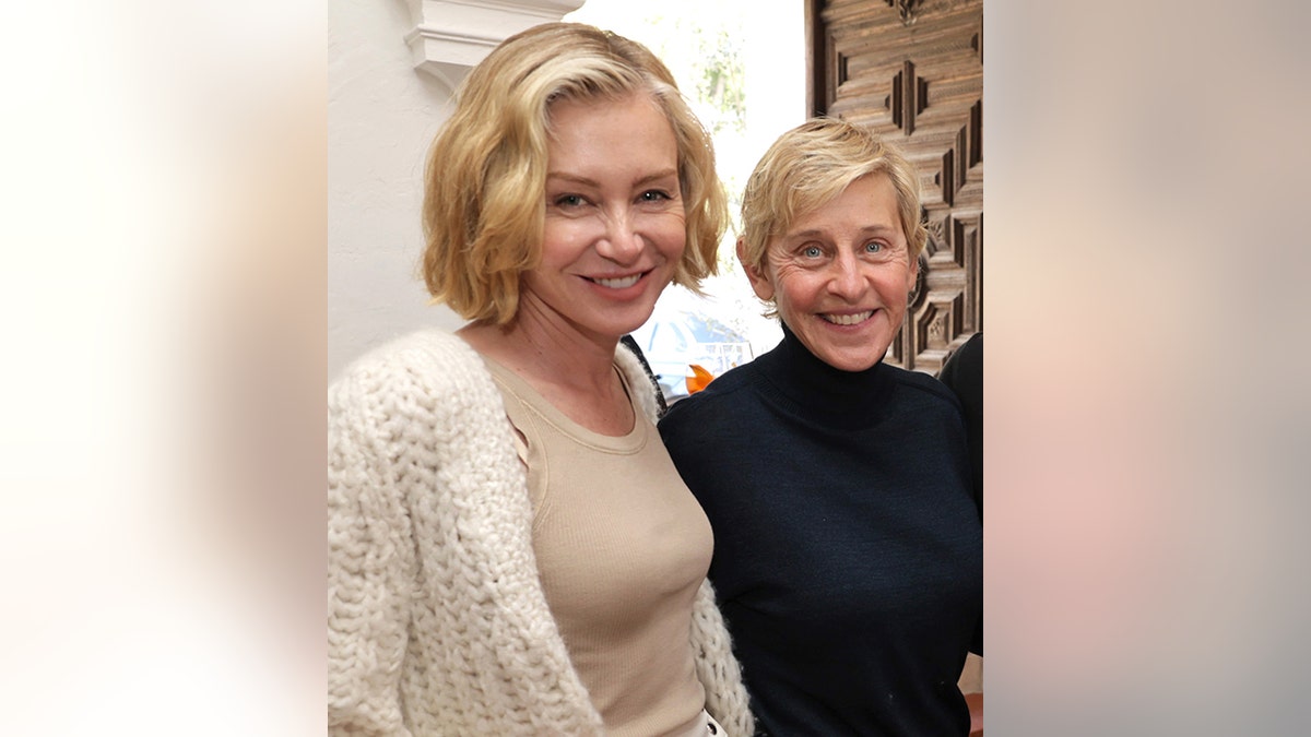 Portia de Rossi in a cream sweater stands with wife Ellen DeGeneres in black