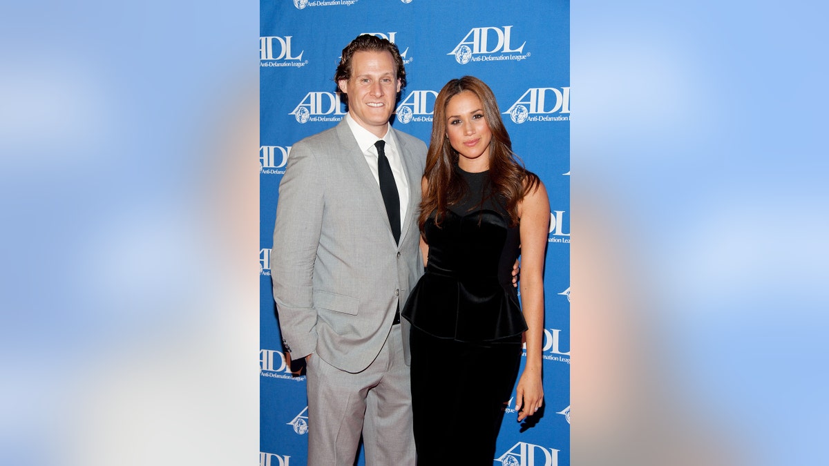 Meghan Markle in a black sleeveless dress standing next to Trevor Engelson in a grey suit