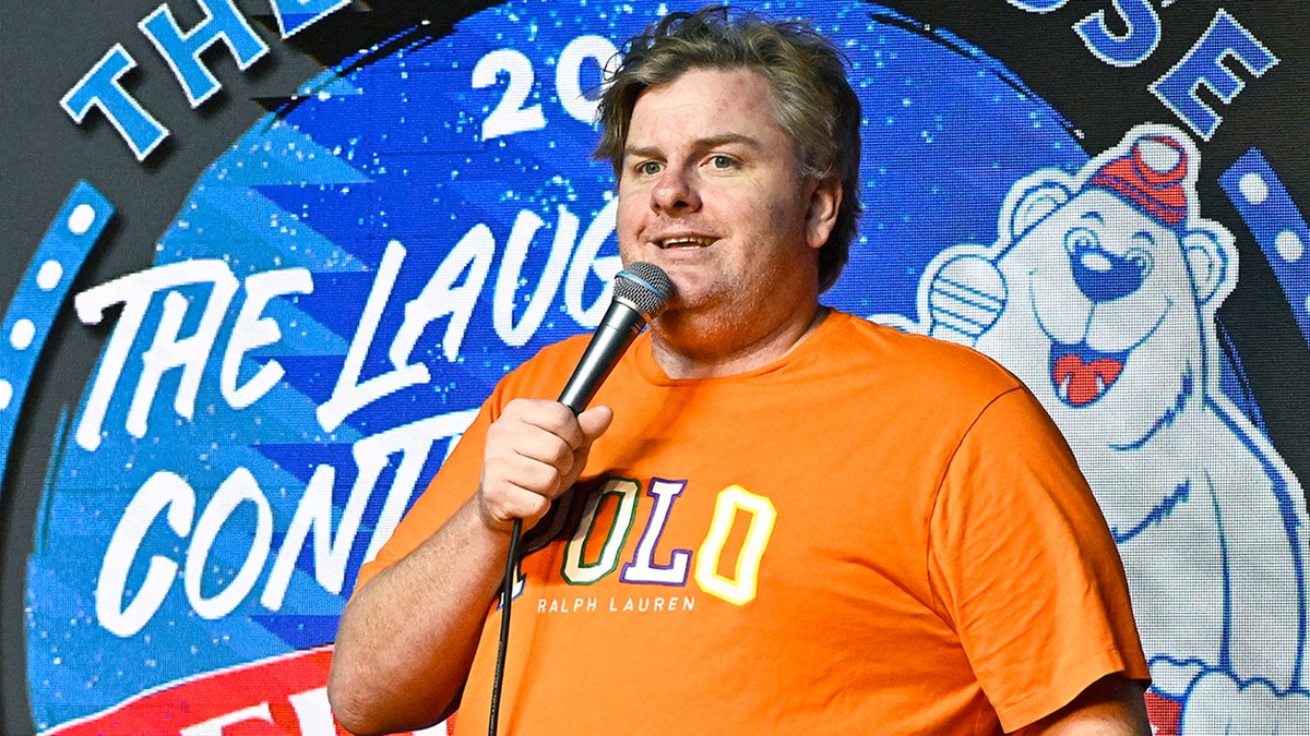 Tim Dillon in a n orange t-shirt that reads 'Polo' stands on stage holding a microphone