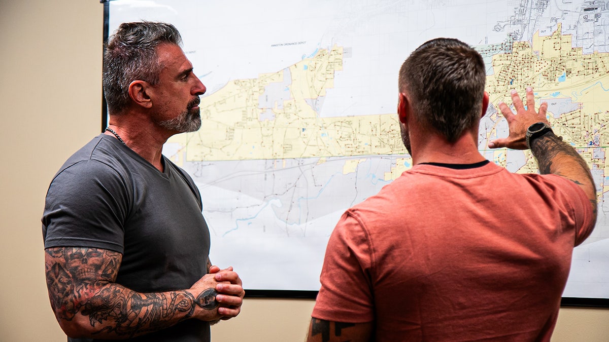 Lou Valoze mirando un mapa y hablando con un oficial vestido de paisano.