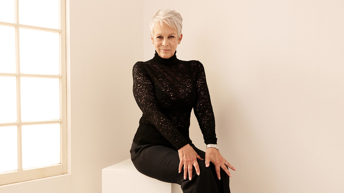 Jamie Lee Curtis sentada en una caja vestida toda de negro y apoyando las manos en las rodillas