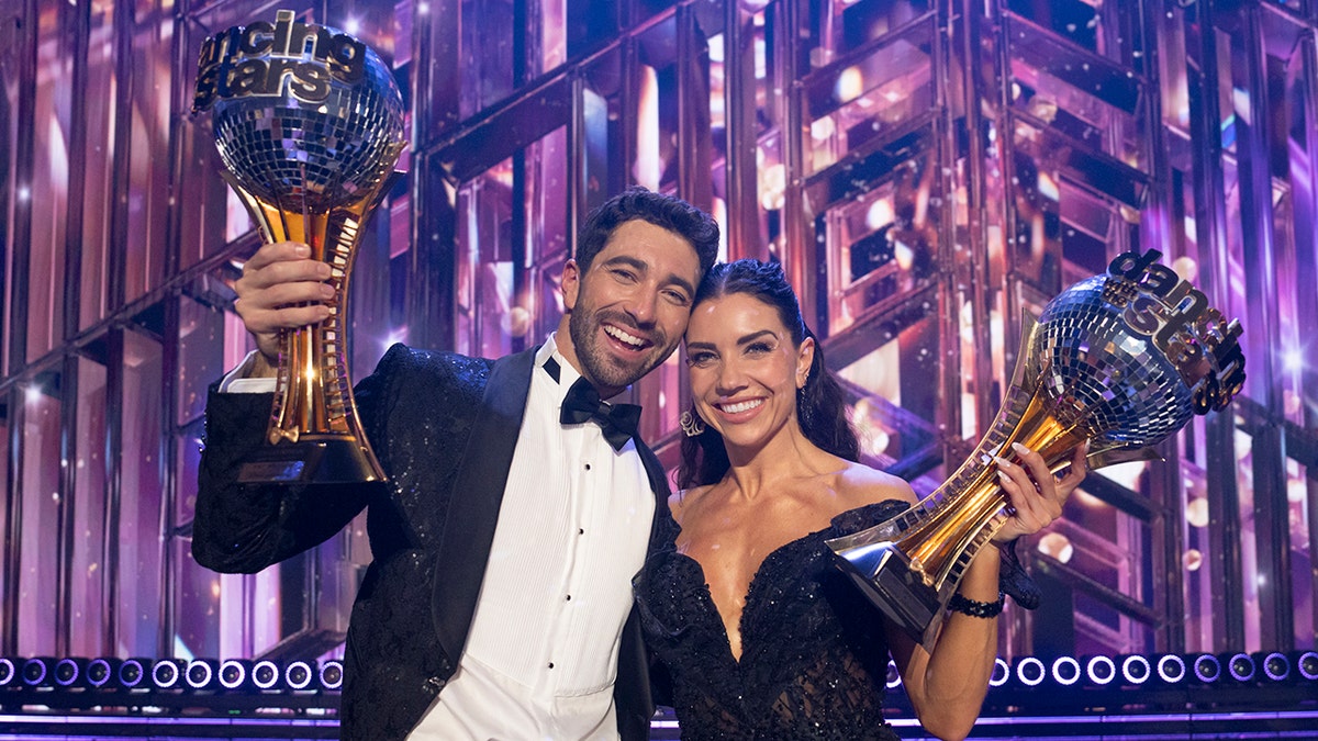 Joey Graziadei con esmoquin y Jenna Johnson con un vestido negro sin hombros sostienen los trofeos Mirrorball en el aire