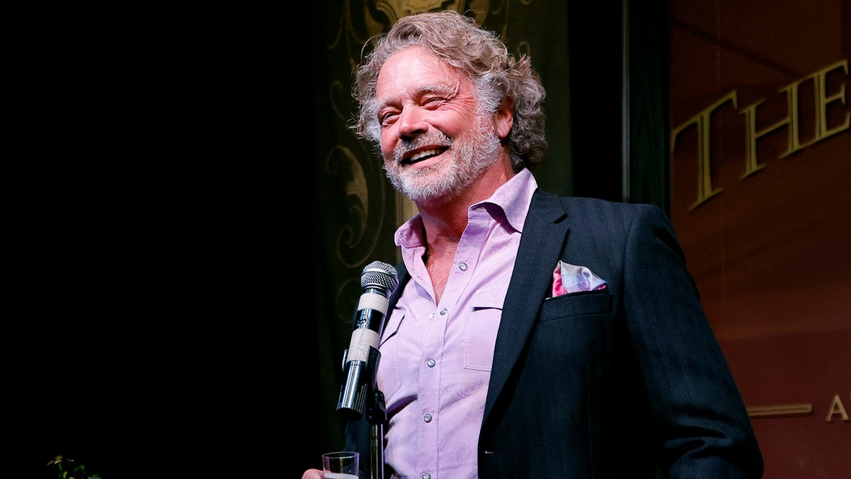 John Schneider in a purple shirt and black suit stands at a microphone