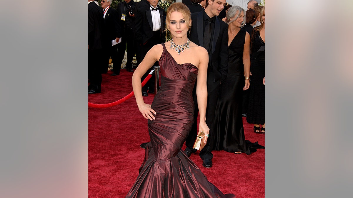 Keira Knightley in a one shoulder maroon, shimmery dress puts her hand on her right hip on the Oscars carpet