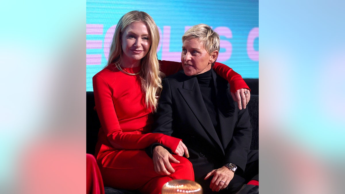 Portia de Rossi in a red dress puts her arm aorund wife Ellen DeGeneres in a black suit