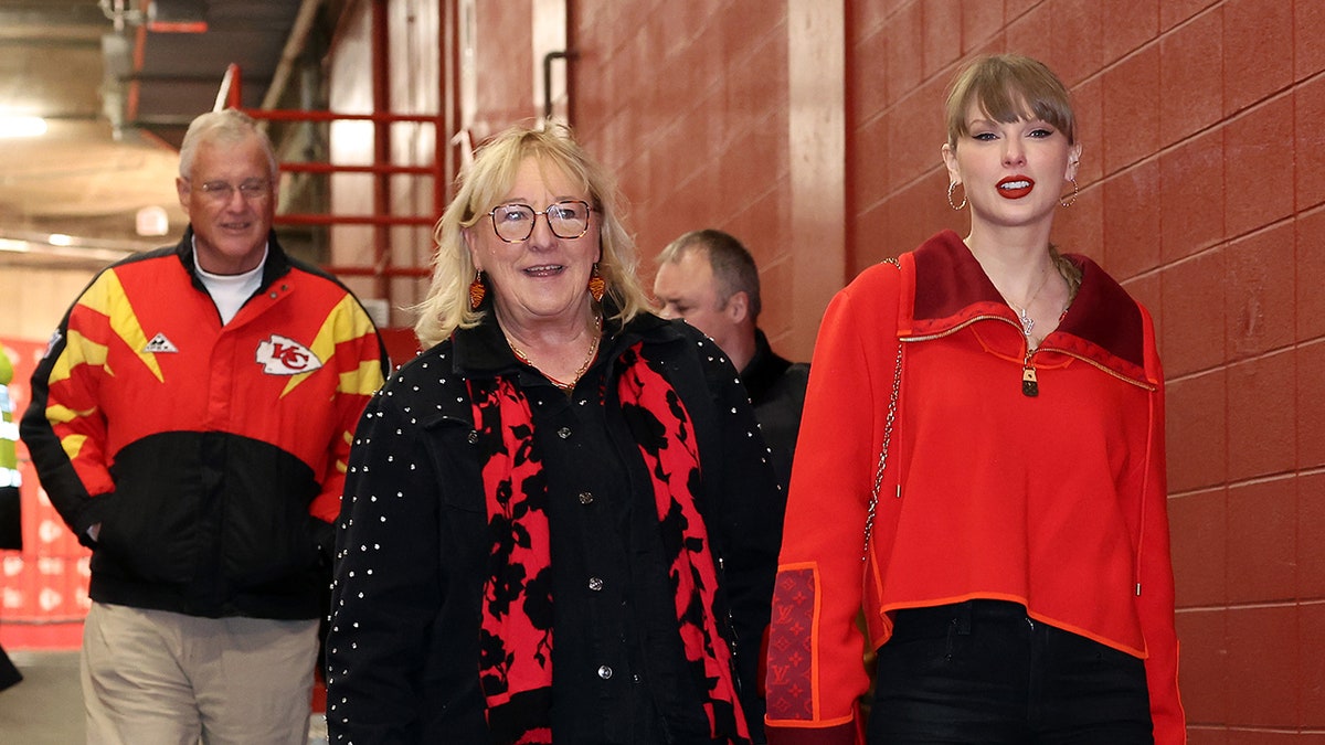 Taylor Swift in a red jacket walks in front of Donna Kelce in a black jacket with red designs in front of Scott Swift in a Kansas City Chiefs jacket