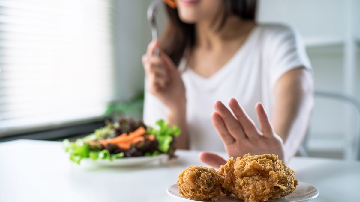 A woman eats a salad and pushes a plate of fried chicken.