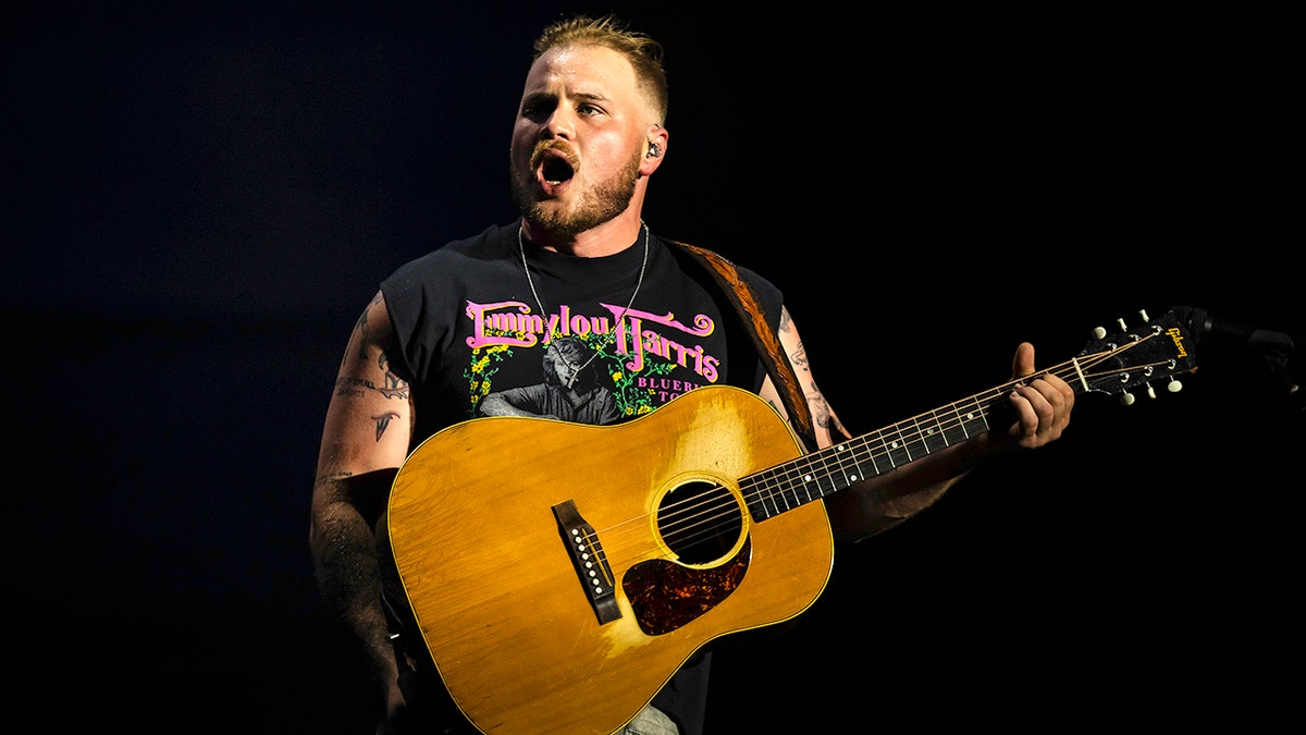 Zach Bryan is wearing a black cropped t-shirt and looks a bit animated/angry on stage with his mouth open with a guitar