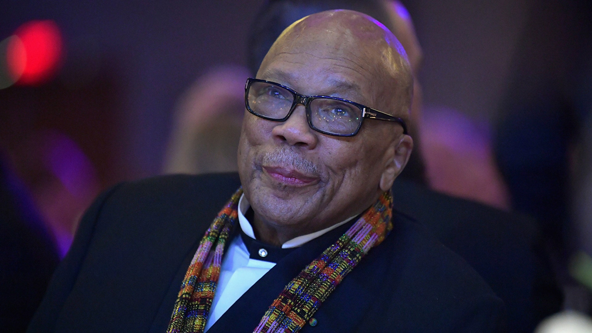 Quincy Jones soft smiles and looks up wearing a suit and colored scarf