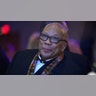 Quincy Jones soft smiles and looks up wearing a suit and colored scarf