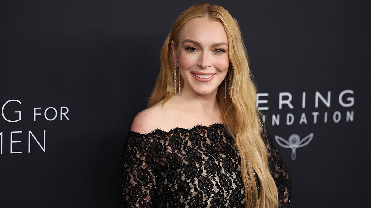 Lindsay Lohan in an off-the shoulder lacy black outfit smiles on the carpet