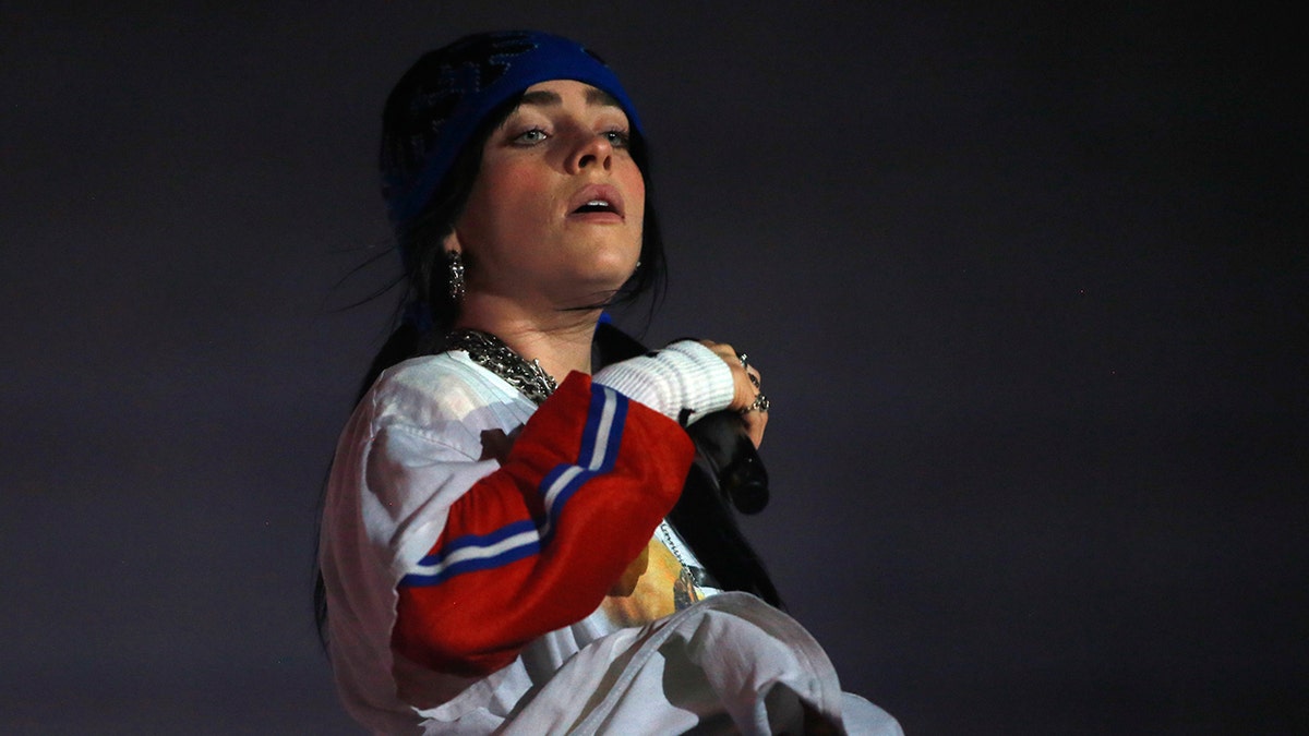 Billie Eilish performing on stage, wearing a blue beanie and multicolored long-sleeved shirt, with her head slightly tilted back.