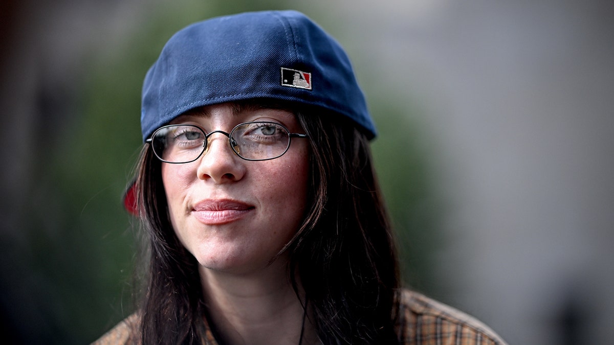 Billie Eilish wearing a backwards baseball hat, glasses, and a soft smile.