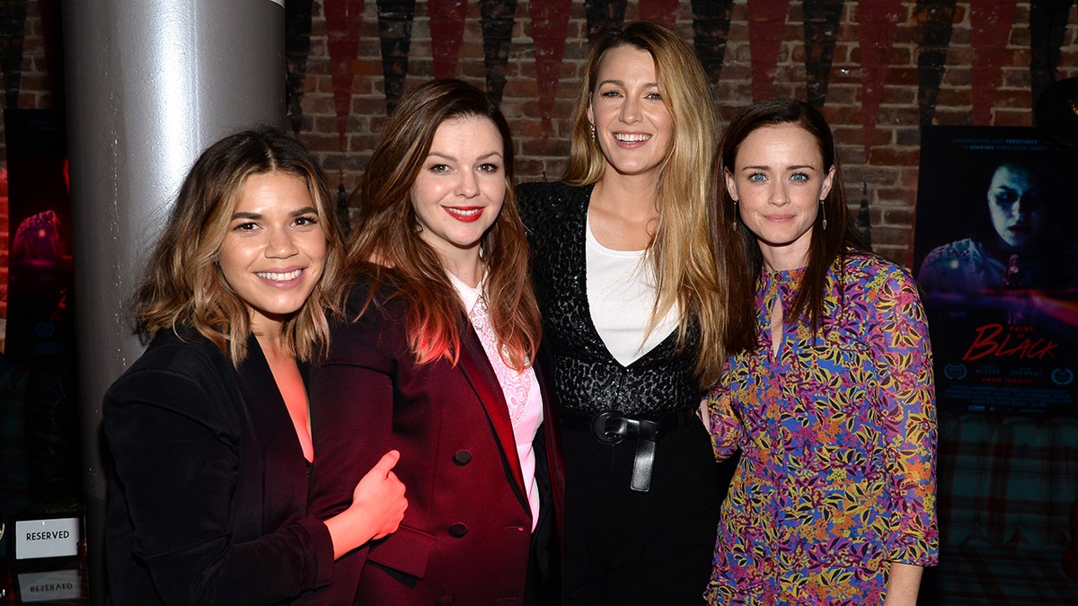 America Ferrera in a black jacket holds onto Amber Tamblyns arm, wearing a red suit jacket and white lace top stands next to Blake Lively in a black sweater top over a white shirt next to Alexis Bledel in a printed dress