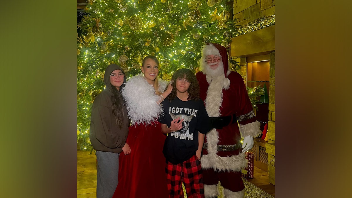 Mariah Carey smiles in a red velvet dress with a white sweater next to her children Monroe and son Morocco, who stands next to Santa Clause