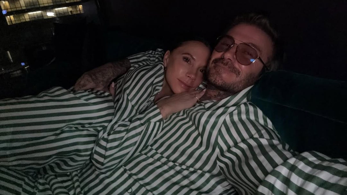 Victoria Beckham in green and white striped pajamas lays with her hand on David Beckham's chest, wearing matching pajamas