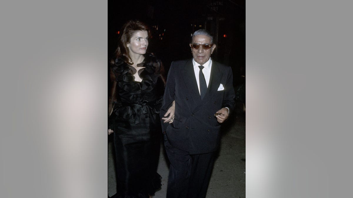 Jackie Kennedy, wearing a coat, held the arm of Onassis, who was wearing a suit.