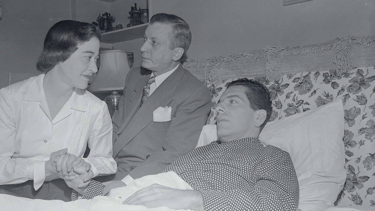 Jennings Lang allongé dans son lit, consolé par une infirmière sous le regard d'un homme en costume.