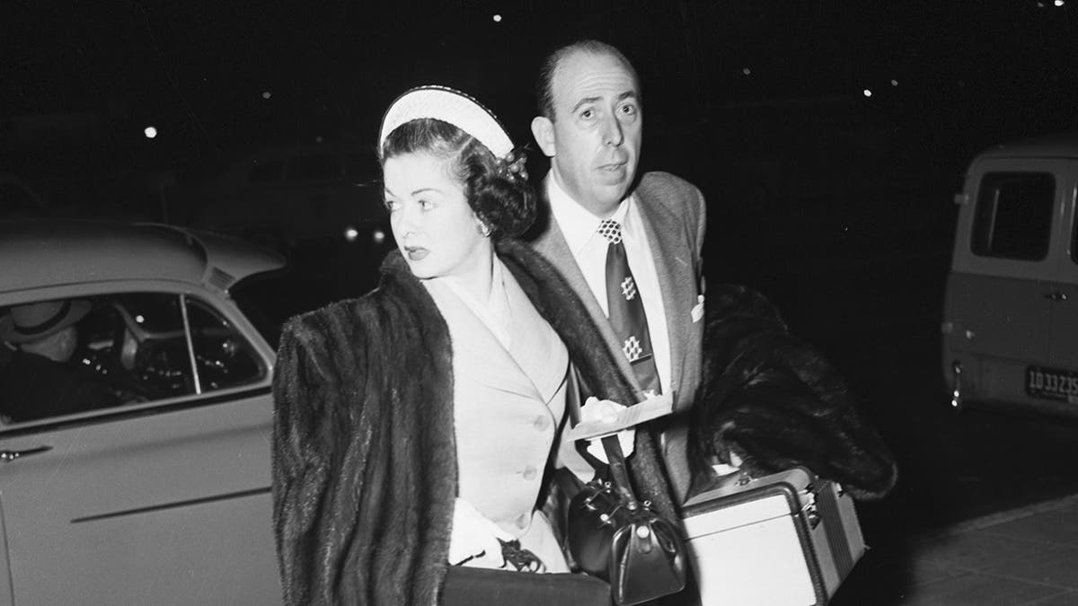 Un gros plan de Joan Bennett marchant dans un manteau de fourrure et un costume à côté d'un homme en costume.