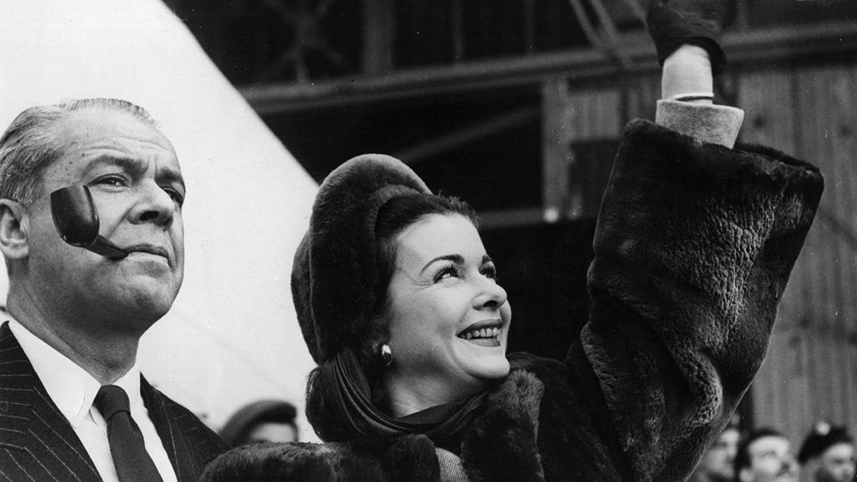 Walter Wanger fumant une pipe à côté de sa femme Joan Bennett en lui faisant signe.