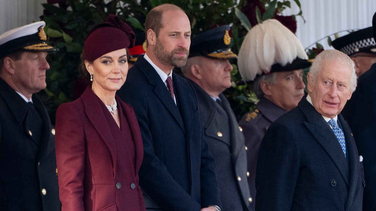 Kate Middleton in a burgundy coat standing next to Prince William and Prince Charles in suits.