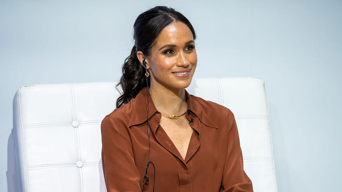 Meghan Markle is sitting on a white sofa wearing a brown blouse.