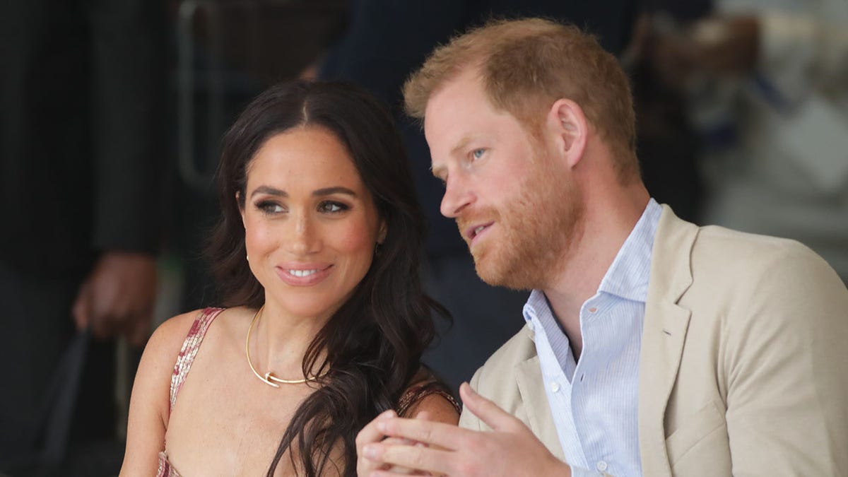 Meghan Markle smiles Prince Harry leaning against her and says something, wearing a blue button down and a sand collar jacket