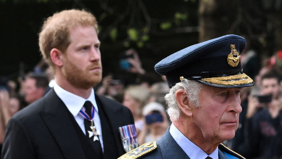 Prince Harry in a suit walking behind his father who is in uniform looking somber