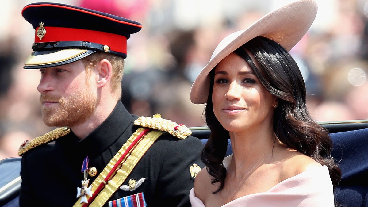 Close-up of Prince Harry in uniform and Meghan Markle in a pale pink dress wearing a hat sitting in a royal carriage.