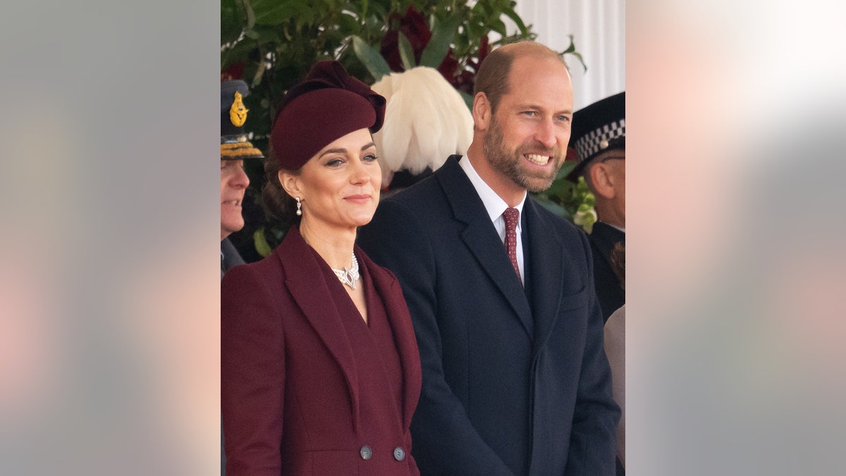 Kate Middleton in a burgundy coatdress standing next to Prince William in a suit and matching tie and smiling
