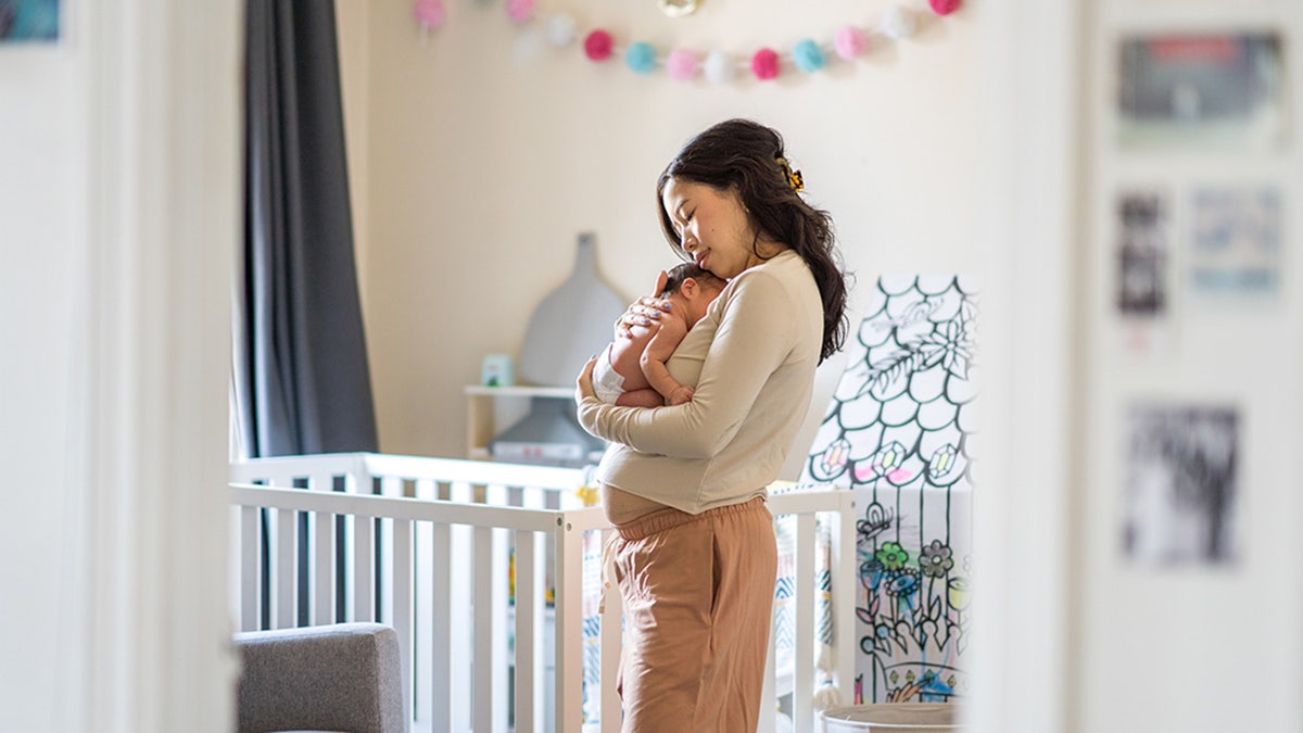 A new mother stands in her daughters' nursery, holding her baby