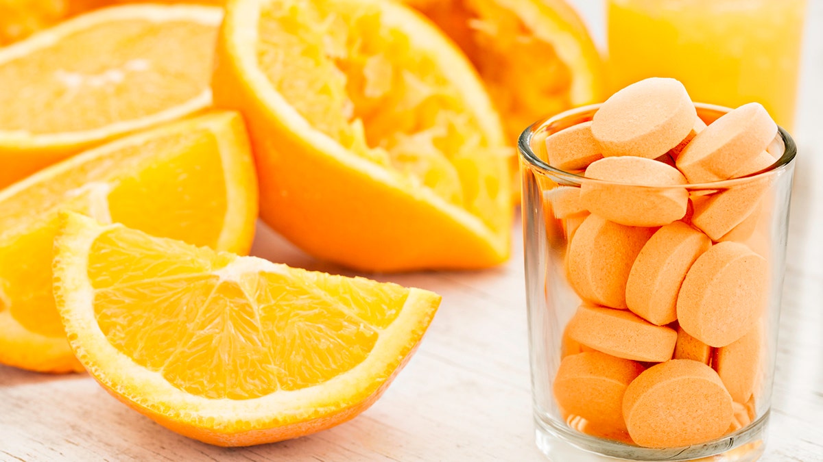 A close up shot of sliced and squeezed oranges a glass of orange juice and a glass full of orange flavored vitamin C Pills