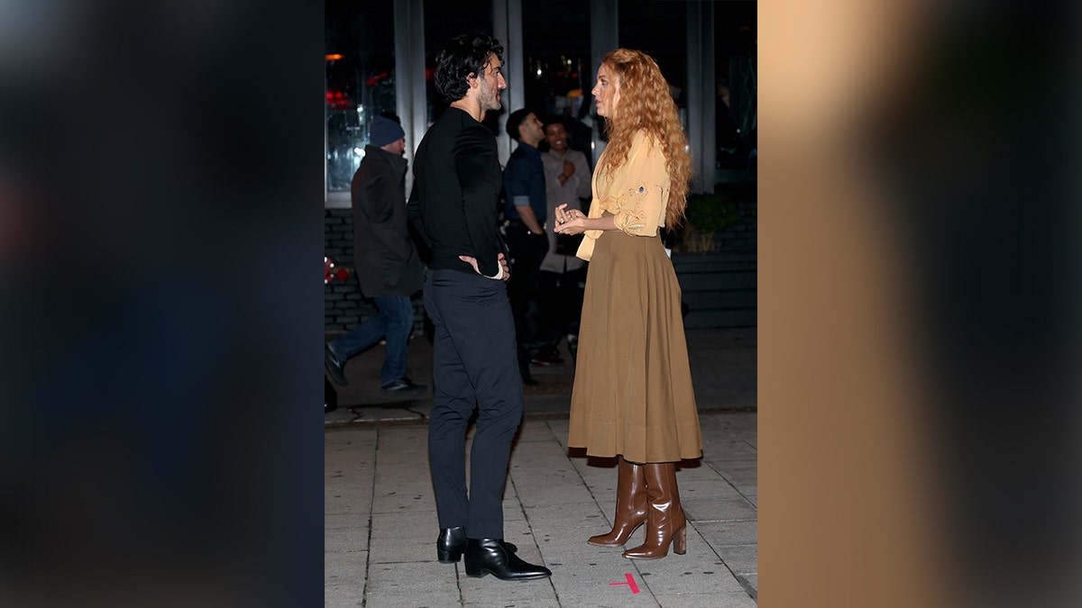 Justin Baldoni in black shirt and navy pants chatting with Blake Lively in orange jacket and brown skirt on a movie set 