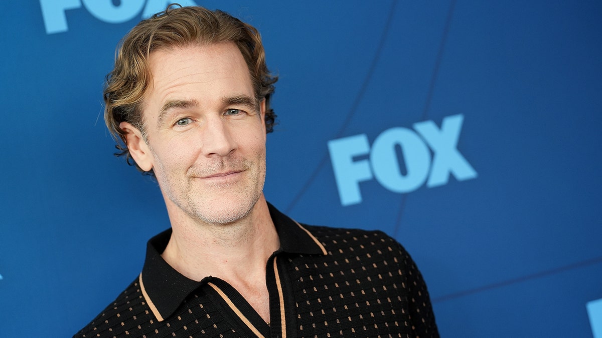 James Van Der Beek in a black fabric collared shirt with brown trim soft smiles on carpet