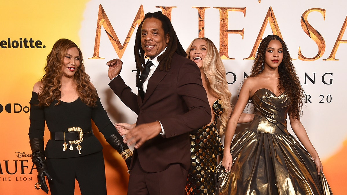 Jay-Z con un traje granate sonríe mientras camina delante de su esposa Beyoncé con un vestido dorado que se ríe y su hija Blue Ivy con un vestido dorado y su suegra Tina Knowles con un traje pantalón negro