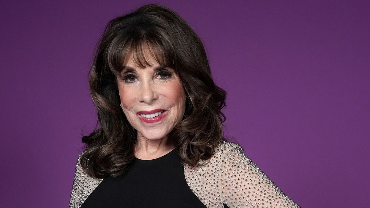 Kate Linder in front of a purple backdrop smiles for a photo wearing a black dress with sequined sleeves