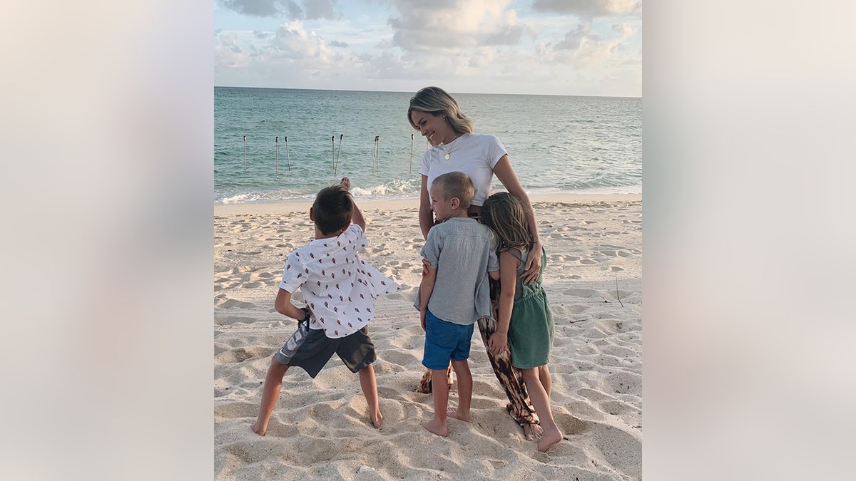 Kristin Cavallari, con una camiseta blanca, sonríe mientras observa a su hijo bailar en la playa, mientras sus otros dos hijos se apoyan en ella