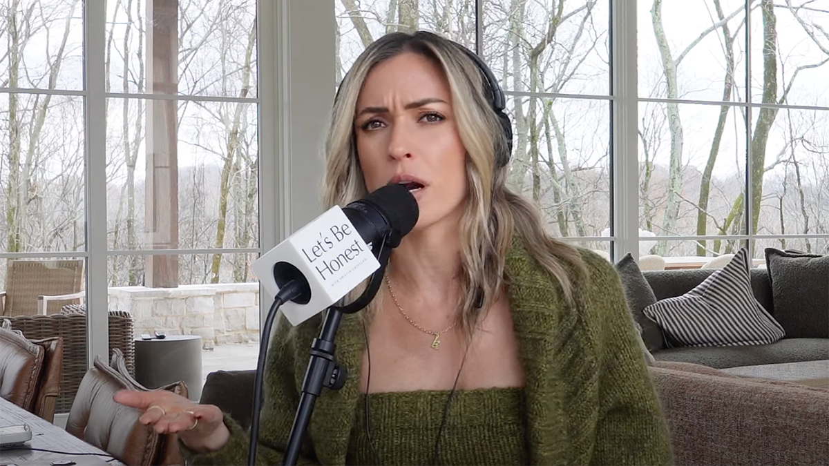 Kristin Cavallari con un conjunto de jersey verde olive mira directamente a la cámara mientras habla en su podcast, llevando unos grandes auriculares negros