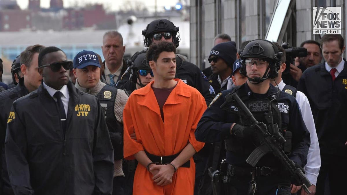 Mangione in an orange jumpsuit and shackles escorted by a large group of tactical NYPD officers