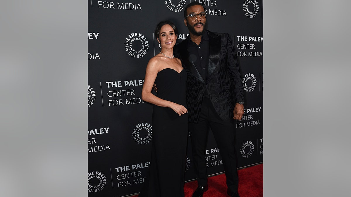 Meghan Markle in a black strapless gown smiles next to Tyler Perry in an all black suit at the Paley Honors carpet