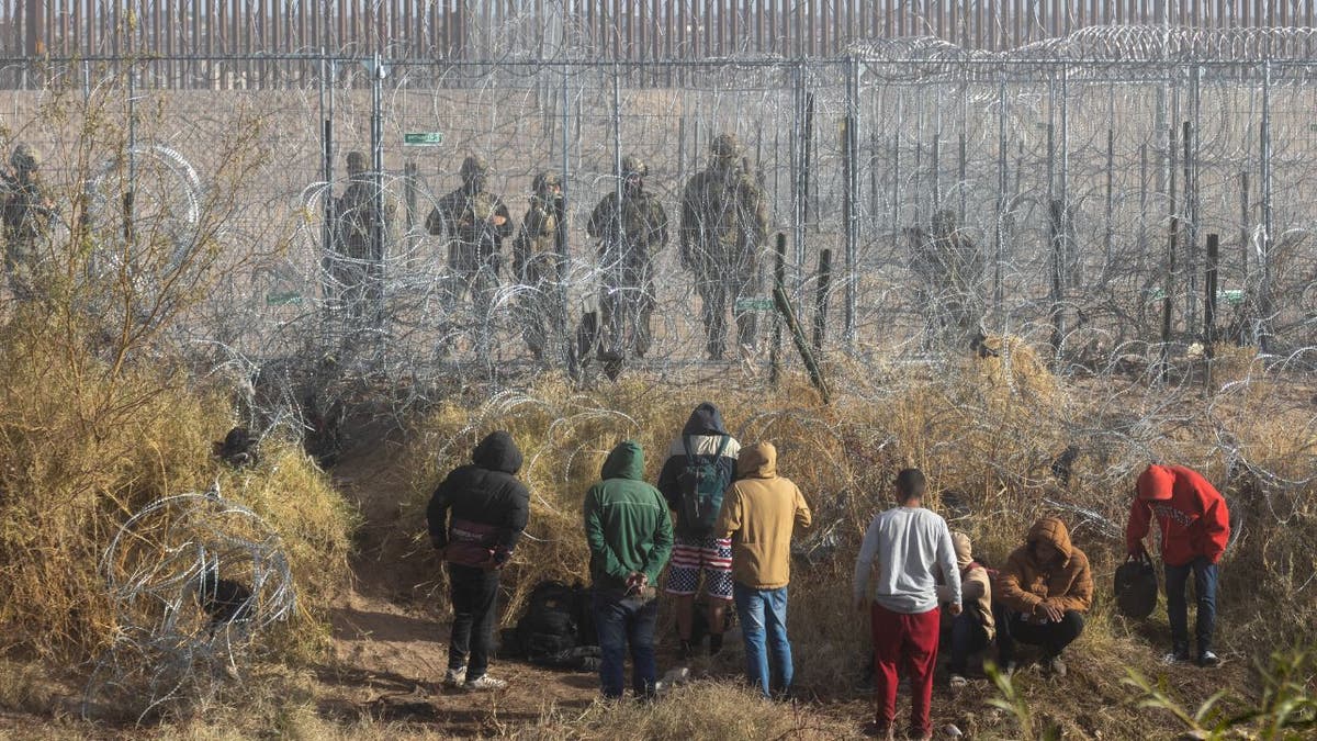 migrants at border, agents on other side of fencing