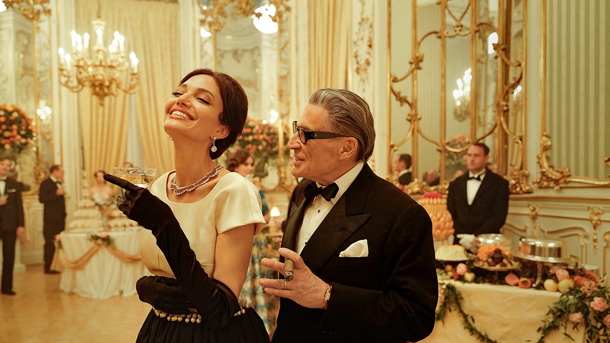 Angelina Jolie smiles in a glamorous gown as Maria Callas, flanked by a suit-clad actor as Aristotle Onassis