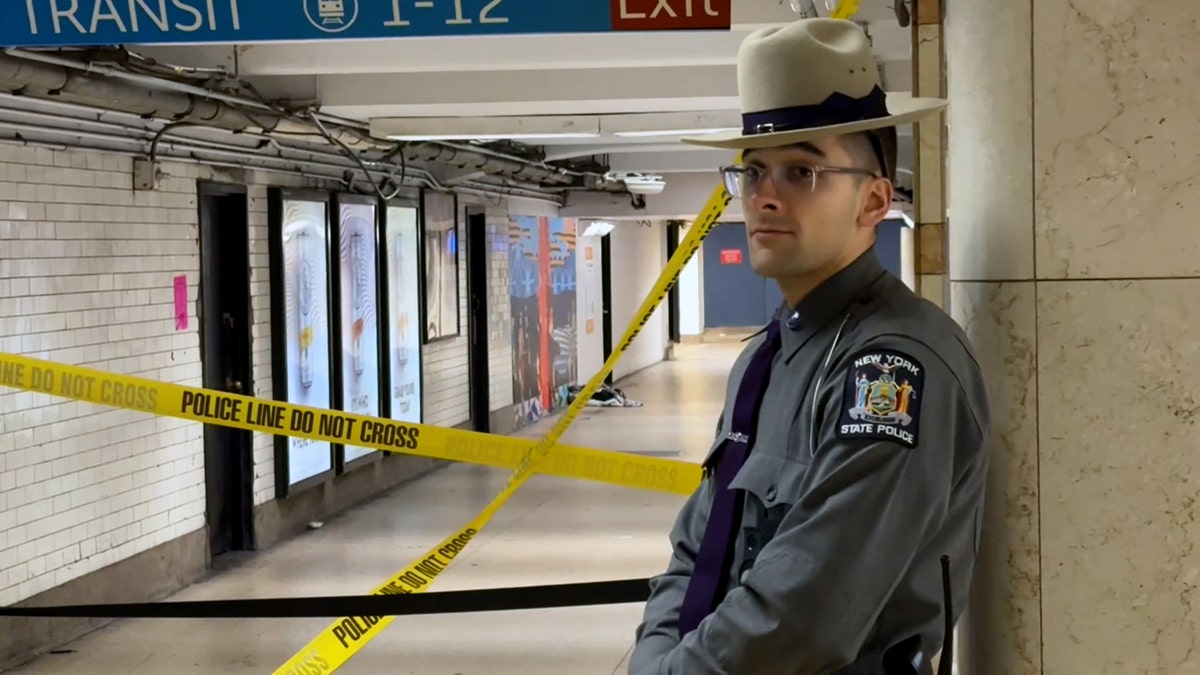 A New York State police officer wearing glasses, a hat, and a gray uniform stands guard outside of a Penn Station hallway cordoned off with caution tape.