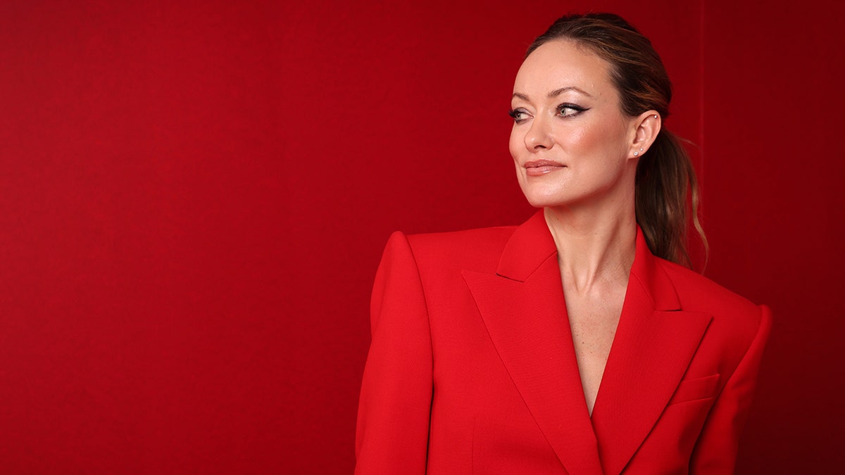 Olivia Wilde in a red blazer looks to her right and shows her side in front of a red background