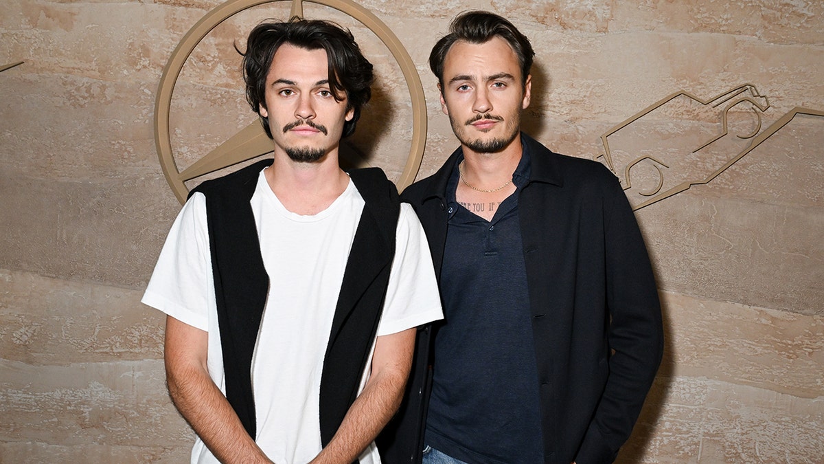 Dylan Lee con camisa blanca y chaleco fino negro y Brandon Lee con chaqueta negra y camisa azul marino en la alfombra