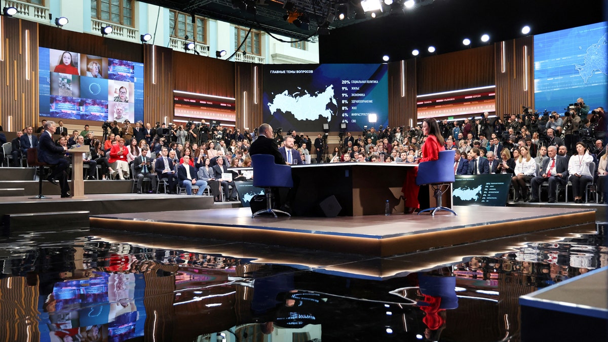 People attend Russian President Vladimir Putin's annual televised year-end press conference and phone-in held in Moscow, Russia. Putin sits at a desk on a central, square stage. Attendees sit in chairs surrounding it.