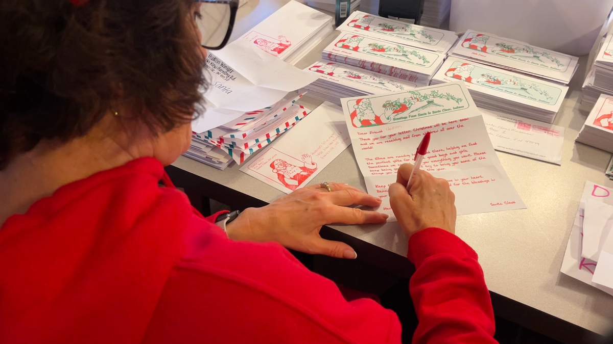 A volunteer in a red sweater writes a reply to a child’s letter to Santa Claus, surrounded by stacks of festive stationery and envelopes.