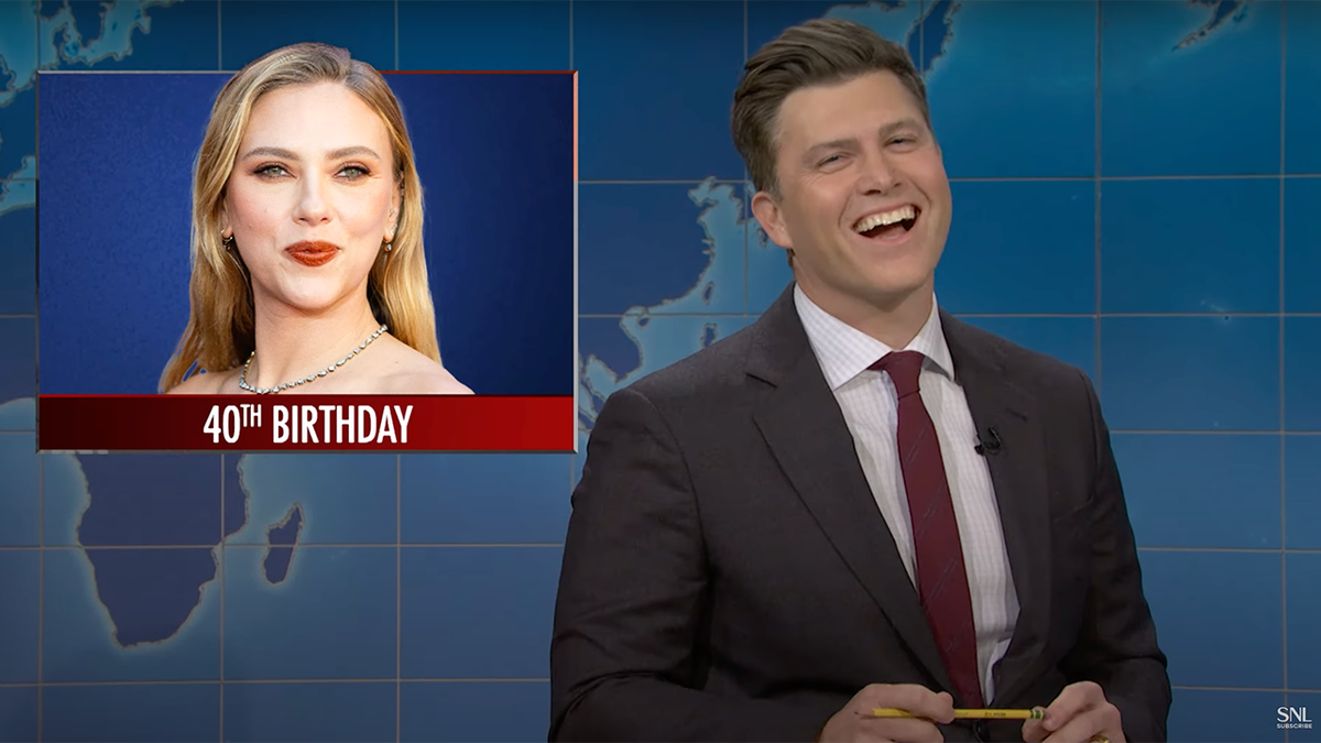 Colin Jost in gray suit and red tie laughing from behind "saturday night live" 'Weekend update" There is a picture of Scarlett Johansson on the table with the words "40th birthday'