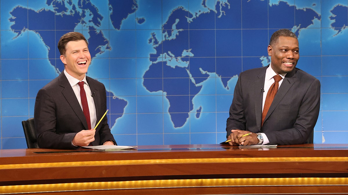 Colin Jost laughs out loud holding a pencil behind the 'Weekend Update' desk on "SNL," wearing a dark grey suit and red tie sitting opposite Michael Che in a dark grey suit and orange tie looking off into the audience, smiling