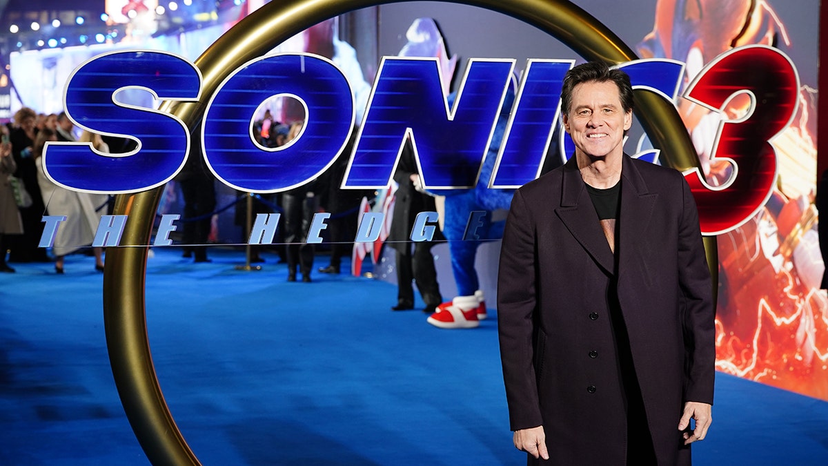 Jim Carrey, wearing a black jacket, smiles in front of a 'Sonic the Hedgehog 3' sign